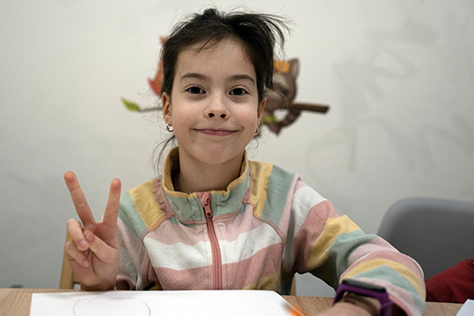 A photo of a young girl from Ukraine attending a therapeutic art class as she smiles at the camera and holds up two fingers in a peace sign.