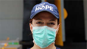 A Project HOPE health worker wears a navy cap with our logo and a medical mask as she stares straight ahead at the camera with dark brown eyes and red hair.