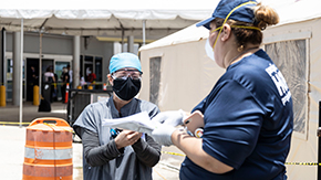 A Project HOPE team member speaks to a doctor in blue scrubs as she holds a clipboard listing supplies to distribute. They both wear masks and head coverings.