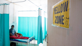 A photograph of a light brown door in India features a yellow sign, reading, YELLOW ZONE: 0 beds. The door is halfway open. Inside you can see the silhouette of a patient sitting on a hospital bed with a breathing mask and red blanket.