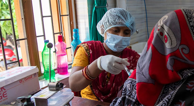 A health worker in India looks down at the arm of a patient wearing a red and gray headscarf as she provides a COVID-19 vaccination. The health worker wears a blue PPE mask, gloves and cap with brightly colored bottles and medical supplies in the background.