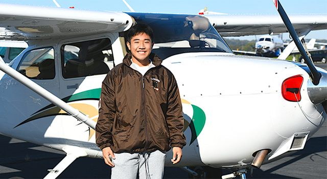 A young man in a brown reflective jacket and gray pants smiles brightly at the camera as he stands in front of a small plane with a front propeller, painted with a green and gold swoosh.