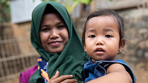 A woman in Indonesia is wearing a forest green headscarf and maroon scarf as she smiles and holds her young child to her chest. The small girl has a curious look and wears a denim jumper.