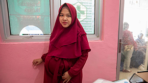 A young pregnant woman in Indonesia is wearing a maroon headscarf and dress as she smiles and holds her stomach and the wall in a hospital. She stands in front of a salmon pink wall and waiting room window with a green medical cross decal.