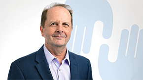 Project HOPE's Chief Health Officer, Tom Kenyon, smiles in a navy suit against a background of two blue hand icons that make up part of Project HOPE's logo.