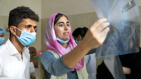 A photo of a doctor looking at x-rays which she holds up to the light. She is wearing a pink headscarf and a blue medical mask.