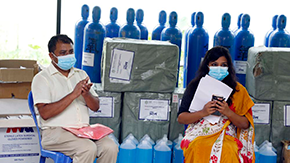 A man in khaki pants and a woman in an orange sari are wearing blue medical face masks, against a background of blue oxygen tanks and other medical supplies in boxes. The woman is holding a pile of papers in her arms and her cellphone in her hands.