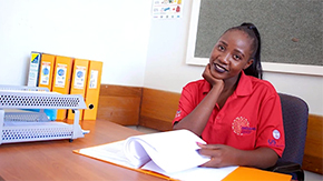 A young woman sits at a desk with her hand holding up her face as she pages through a binder of papers. She has long black hair and wears a red polo as she smiles brightly in Namibia.