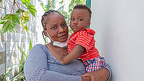 A photograph of a  mother holding her baby in Haiti. The mother has braided hair and a slight smile as her mask is pulled down. She wears a gray shirt, and her baby wears a red polo and plaid shorts. They stand against two gray walls outiside as plants and vines hang down.