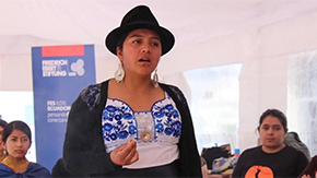 A young woman of the Kañari People of the Kichwa Nationality of Ecuador is wearing a black hat and sweater with a white and blue embroidered blouse. She is emoting with her hands as she speaks to a classroom of her peers and students.