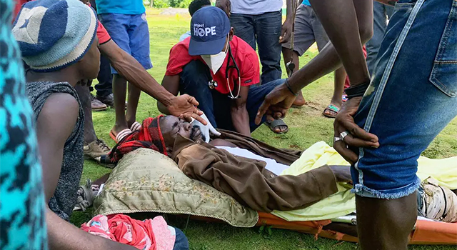 A man in a red shirt and blue Project HOPE hat and medical mask is examining a man lying prone on a stretcher as other people gather around offering help. The man was dug out of the rubble after a landslide caused by the combination of the earthquake and tropical depression.