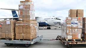 A commercial airplane is the background of this photograph of two huge pallets of supplies with boxes piled high. They are labeled with the Project HOPE label.