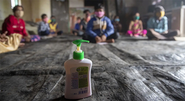 A photograph of a classroom with a bottle of hand sanitizer in focus in the middle. A group of children sit in the background just out of focus.