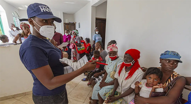 A photograph of a Project HOPE health worker in navy blue t-shirt and cap talks to women sitting in a clinic in Haiti as they wait to receive medical care.