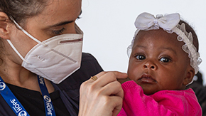 A photograph of a Project HOPE emergency responder holding and playing with a baby in Haiti. The baby wears a pink shirt and white lacy headband with a bow.