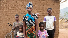 A family in Malawi stand in front of their home. The mother wears a blue head wrap and carries a baby on her back as her four other children stand behind her.