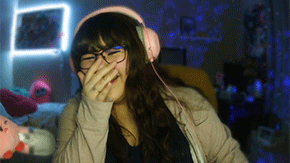 A screenshot shows a young woman with long black hair and bangs covers her face with her hand as she laughs, streaming live. Her eyes are closed and she wear pink headphones, black frame glasses and a tan sweater over her black shirt.