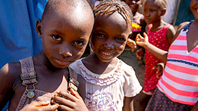 A photograph of two small children playing outside. They smile at the camera as the sun illuminates the top of their faces and shades their lively and curious faces.