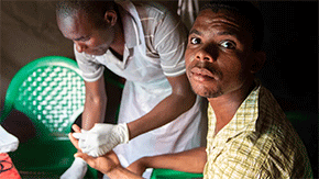 A man looks up at the camera as he receives an injection of his medication in Malawi. He wears a yellow plaid shirt as the man giving him the injection wear gloves and apron over a tan striped polo shirt.