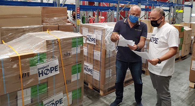 Two men, one wearing a white Project HOPE t-shirt, the other a navy polo. Both inspect paperwork while wearing masks in a warehouse filled with boxes, labelled with the Project HOPE logo and covered in plastic.