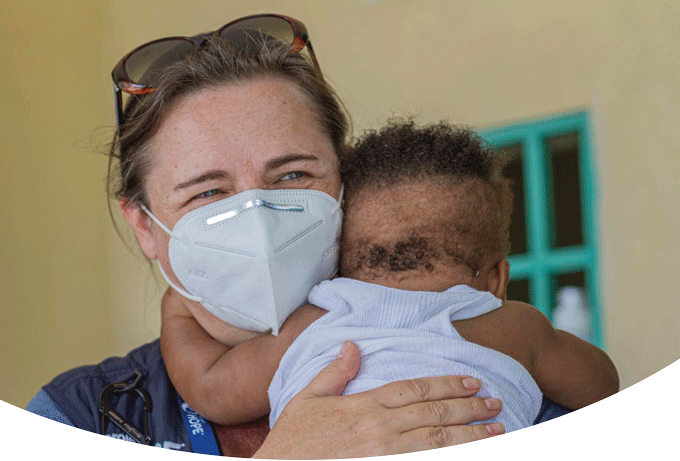 A member of the Project HOPE Emergency Response Team, Cora Nally, wears a facemask, sunglasses and a blue Project HOPE vest, as she holds a small baby wearing a white shirt. The baby has its arms around her neck as Cora smiles with her eyes.