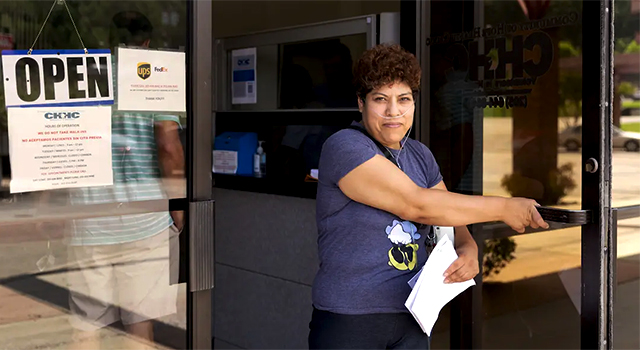 A women with dark curly, short hair is opening up a windowed door of a community clinic. She is wearing a blue tshirt, holding some paperwork and looking at the camera with a slight smile.