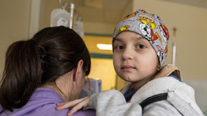 A young child wearing a gray sweatshirt and patterned hat looks at the camera quizzically as her mom, with the back of her head towards the camera looks down, holds her inside a room with medical equipment.