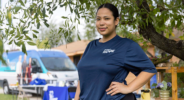 A Project HOPE team member stands with her hand on her hip and smiles. Her hair is black and pulled back. She is standing next to a tree and a small house and van are in the background as she is giving vaccinations in neighborhoods in her navy Project HOPE t-shirt.