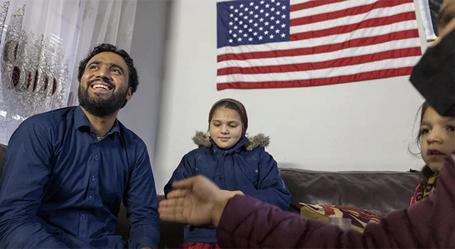 A family of 4 sits under a big fabric American flag. The father of this family from Afghanistan is looking up and smiling while his two children and wife sit nearby.