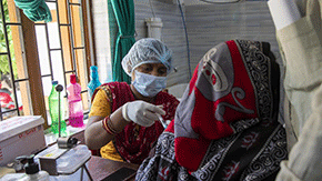 A Project HOPE-funded community health worker in India wears bright red and yellow outfit and PPE including cap, mask and gloves as she gives a vaccine to a woman with a red scarf covering her head.