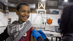 A young boy is smiling as he looks to the right. He is wearing a gray sweatshirt and red tshirt with the sleeves pushed up so a nurse with her back to the camera can prepare to vaccinate him against the background of a school gym.