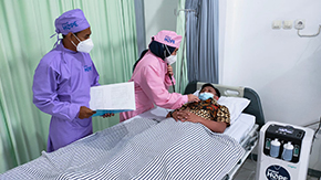 Two Project HOPE-supported health workers in Indonesia help a patient laying in a hospital bed use the oxygen concentrator provided by Project HOPE.