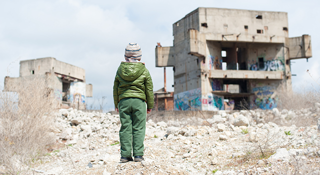 A young child wearing a green quilted jacket, green pants and a gray knit hat stares ahead with his back to the camera. He is looking at the remains of cconcrete buldings in a field of rocks.