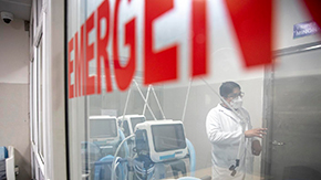 A doctor in glasses, a mask and lab coat is shown talking and extending his hand as he speaks in front of a room of medical equipment. The doctor is shown behind the glass of a room with large red lettering labeling it as EMERGENCY.