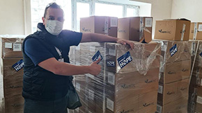 A health worker on the Ukraine border wears navy Project HOPE shirt, jeans and mask as he points to many shipping boxes of medical supplies, set in a sunny room and sent from and labeled with Project HOPE stickers.