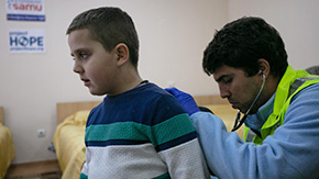 A health professional is a yellow vest, blue shirt and blue surgical gloves is using a stethoscope to listen to the breathing of a young boy refugee from Ukraine who wears a blue and white striped sweater.