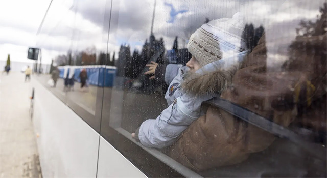 A young child wearing a blue  jacket and tan knit hat stares out the window of a bus leaving the border for safety in Poland. His mother is wearing a brown jacket and holding the child on her lap aboard the crowded bus.