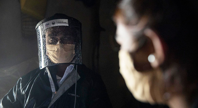 A health care worker wearing a steamed up face shield, face mask and zip up jacket looks to the left in a dark room where they are providing COVID-19 information door-to-door in a community in India.