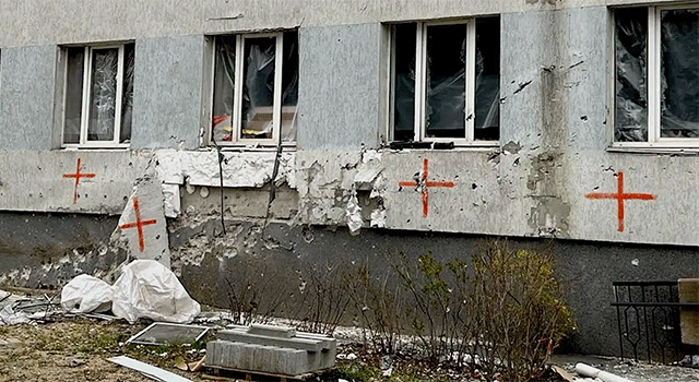 A grey concrete building in Irpin, Ukraine is scarred with bullet and bomb holes and red spray-painted crosses under each window.