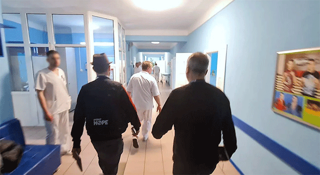 The backs of two men walking down a blue corridor wear dark blue Project HOPE jackets in a hospital in Kyiv.