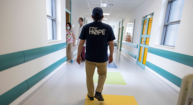 A man in navy blue Project HOPE shirt and cap with khaki pants has his back to the camera as he walks down a white hospital hallway. Two staff in scrubs are walking into a room ahead.