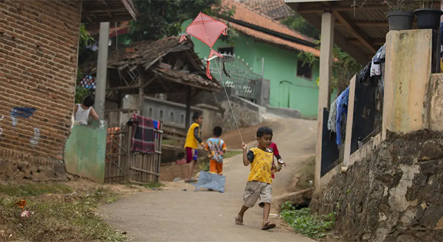 A small boy in India wears a t-shirt, khaki shorts and sandals as he flies a small kite and walks through a village of brick and stone houses.