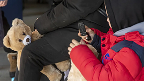 A Ukranian child in a jacket plays with a toy car while waiting for a train to safety. His guardian holds a brown teddy bear on top of their luggage.