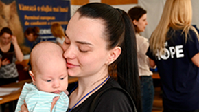 A Ukranian mother holds her baby against her face with a slight smile as the baby looks down. Project HOPE and other organizations' staff talk in the background.