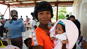 A mother in Haiti wears a hair covering and holds her small baby in a blanket against her chest as she smiles at the camera. She is under a canopy and surrounded by other families and service staff.