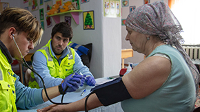 A Project HOPE-sponsored health worker is taking the pulse and blood pressure of a woman and refugee from Ukraine. She wears a scarf on her head, earrings and a blue tanktop.