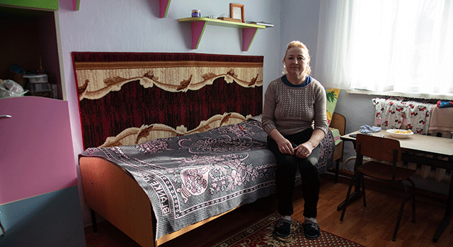 A Ukranian woman with blonde hair in a blue t-shirt, tan sweater and jeans, sits on a patterned spread atop a small wooden bed in a shelter for refugees fleeing the conflict with Russia.