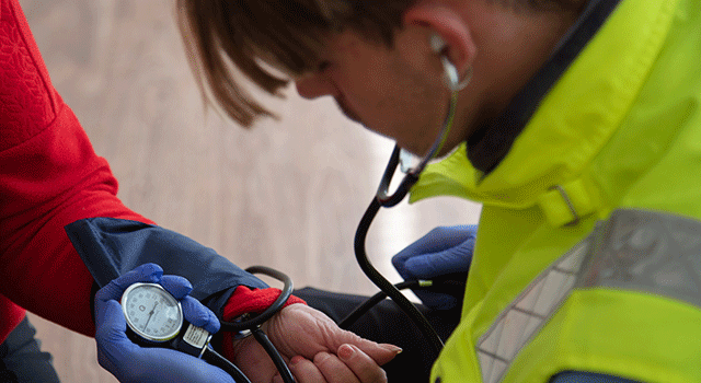 A health care worker in a yellow emergency vest and blue nitrile gloves holds a blood pressure gauge and is looking intently at it as he takes the reading from a person wearing a red sweater. Only their hand is visible.