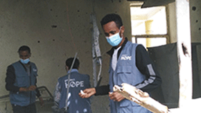 Three health care workers, wearing blue Project HOPE vests and medical face masks, are pictured in a clinc in Ehtiopia holding medical supplies that have been scattered.