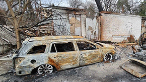 A blown out car sits in front of a mangled building. The trees also show sign of damage from bombing in Ukraine.